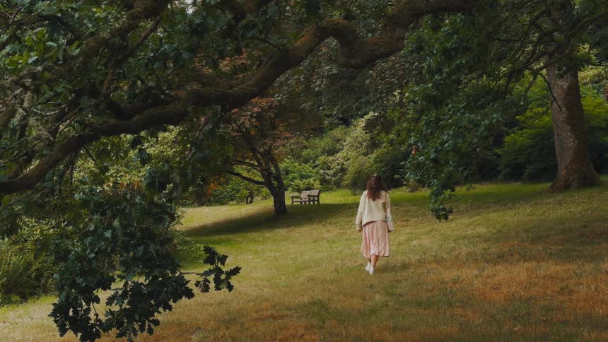 A young woman walking across the park grass at midday heading toward a bench surrounded by trees