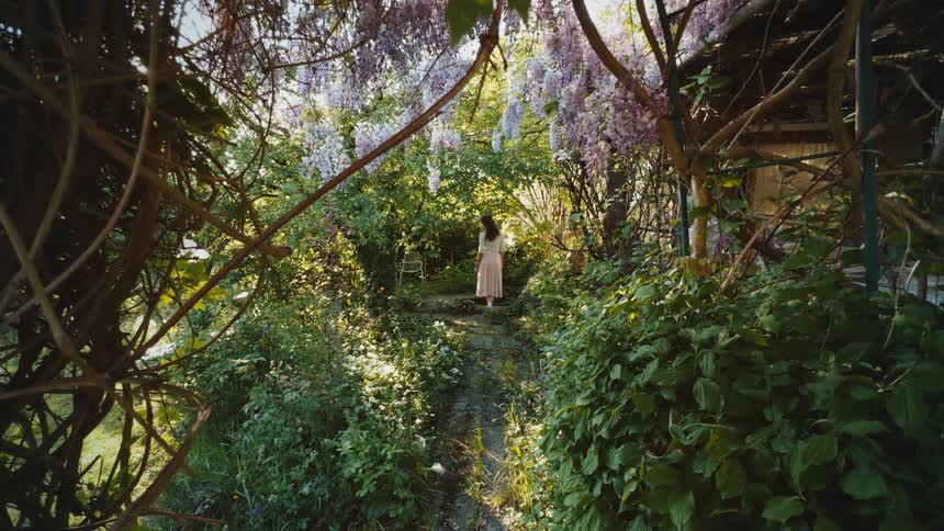 Person walking along a narrow path in a lush garden with dense vegetation and hanging flowers surrounding the scene