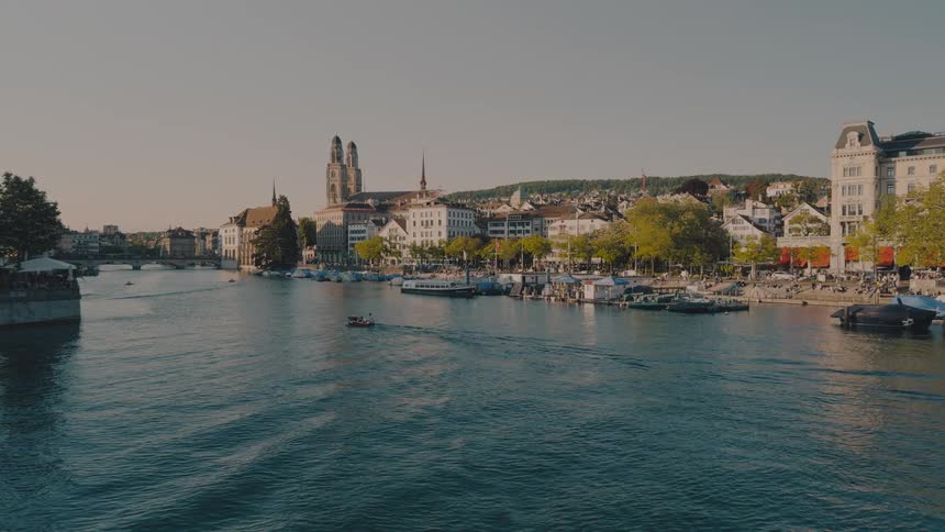 Ship sailing on the water in Zurich as traffic and pedestrians move by buildings on the coast