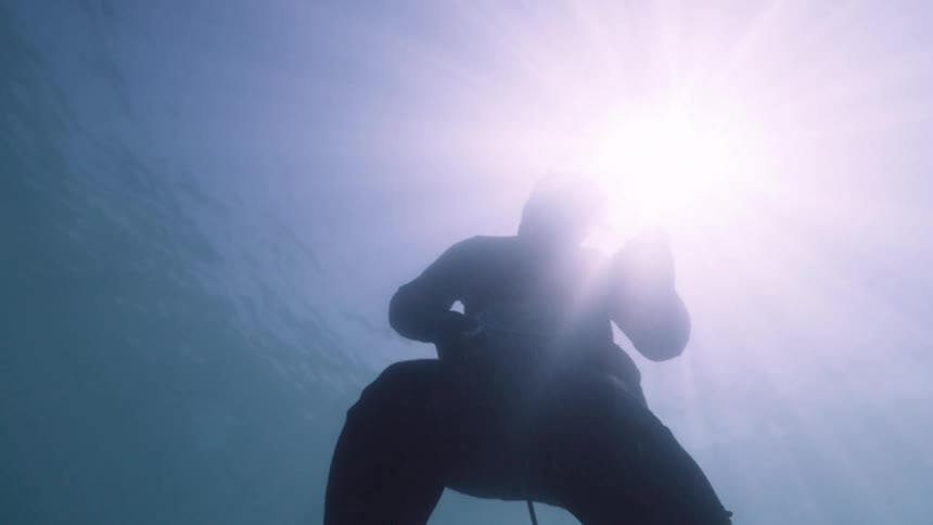 A person wearing a diving mask and a red longsleeved shirt is seen from an underwater lowangle perspective as they swim at the surface of the water with bright sunlight shimmering through from above