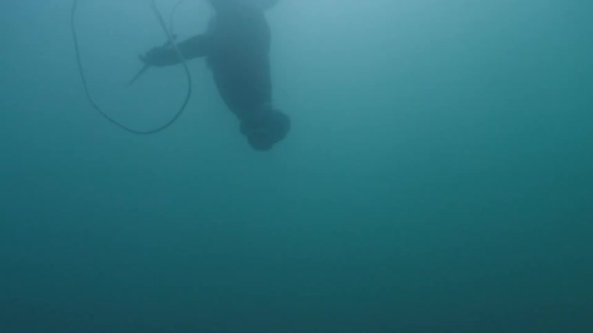 A scuba diver swims headfirst toward the seabed in murky blue water wearing full diving gear and fins while descending past a dark reef formation with a thin rope trailing from above into the deep aquatic environment