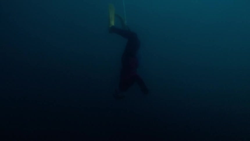 A free diver in a red wetsuit descends vertically into the deep dark blue ocean while following a white guide linethe camera tracks the divers slow controlled movement as they fade into the shadows of the increasing depth