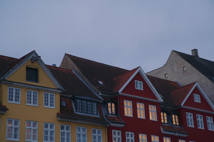 A row of colorful europeanstyle townhouses with steep tiled roofs and glowing windows stands quietly beneath an overcast evening sky creating a cozy livedin street scene