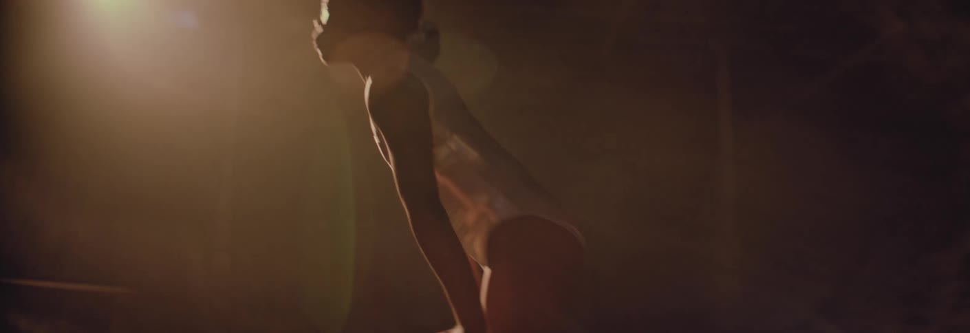 An athletic young gymnast in a white leotard approaches a balance beam and climbs up under a golden spotlight