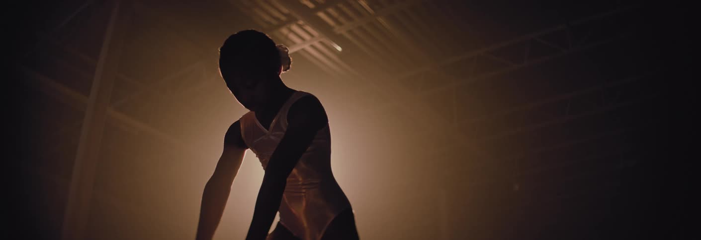 A young female gymnast in a white leotard stands in a hazy backlit gymnasium and practises her finishing stance on the balance beam