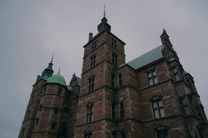 The footage captures a lowangle shot of a grand renaissancestyle castle featuring red brick walls and distinctive green copper spires under a heavy overcast sky the architecture conveys a sense of historical weight and somber elegance