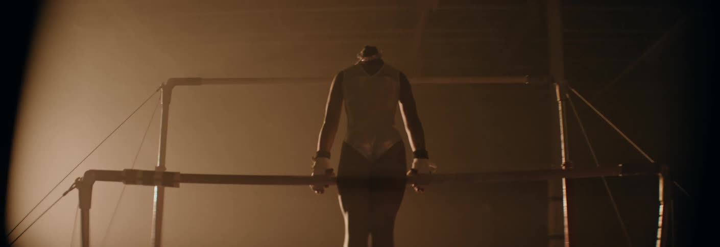 A gymnast in a shimmering silver leotard executes a powerful swing and transition on the uneven bars within a hazy spotlighted gym
