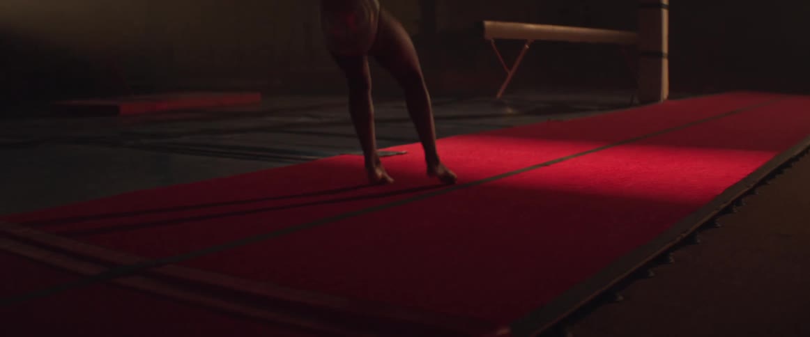 A tracking view of a gymnast performing a series of handsprings across a long red tumbling mat in dark gym