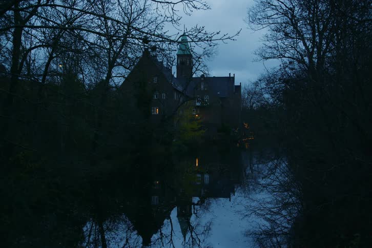 A large building with a greendomed tower sits along a dark river its structure and warm window lights reflecting in the water the scene is framed by bare tree branches under a dim overcast sky at twilight
