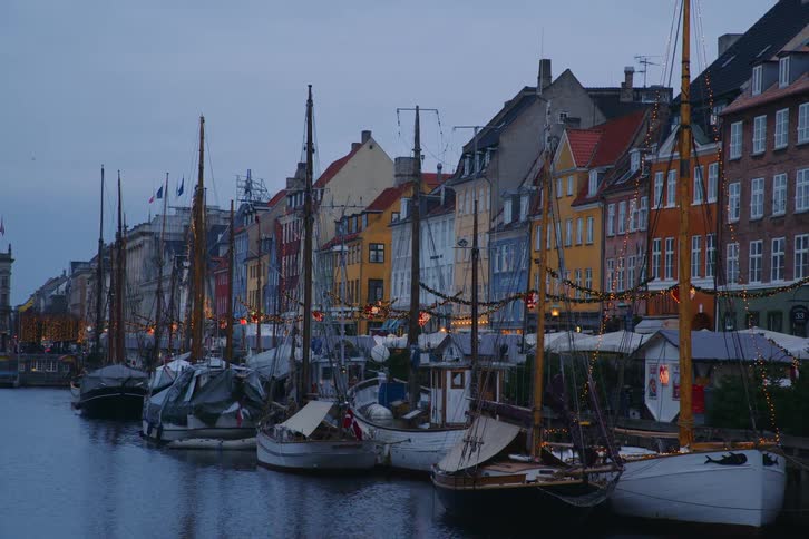 As evening falls over the historic canal colorful merchant houses glow with lights while rows of vintage sailboats rest in the quiet water framed by a distant pedestrian bridge bustling with life and evening traffic