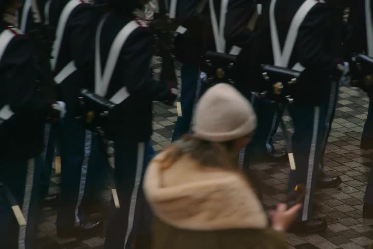 A view from behind shows a group of soldiers in dark blue uniforms with white crossbelts and blue trousers with white stripes marching in unison along a paved street