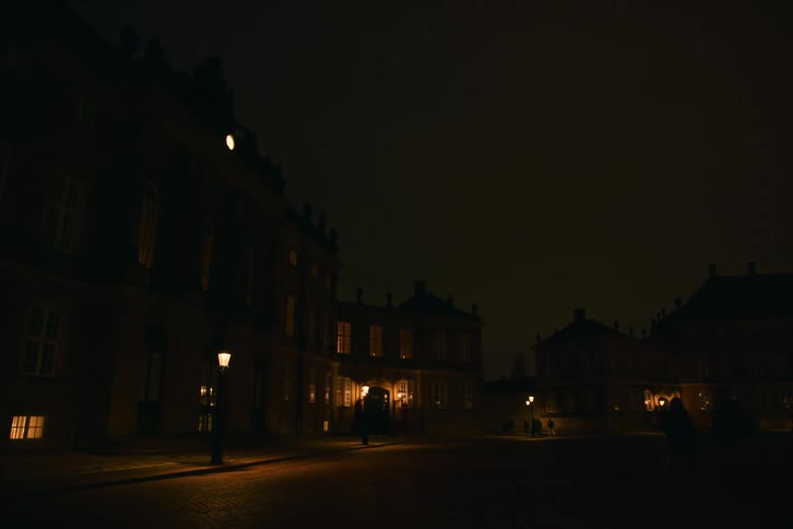 A historic european building complex glows under warm street lamps at night beneath a moody sky