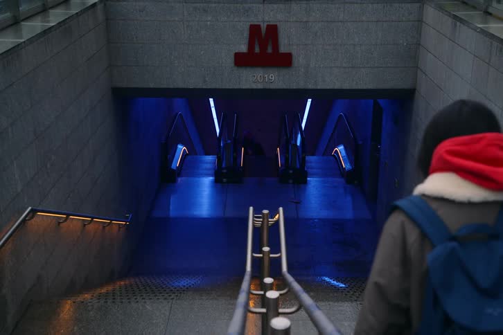 A young woman walking down a staircase into a subway terminal wearing a jacket and a backpack