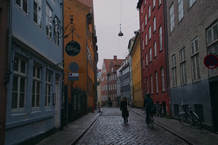 A woman walks down a cobblestone street past colorful european buildings as a cyclist passes