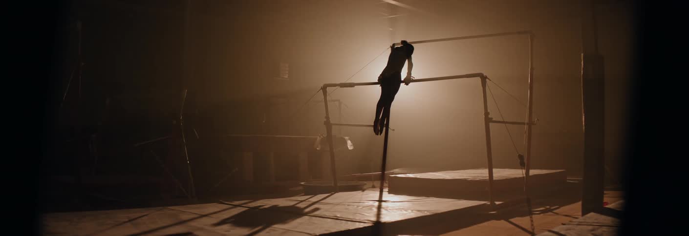 In a dimly lit gymnasium a gymnasts silhouette is captured performing a mount onto the uneven bars