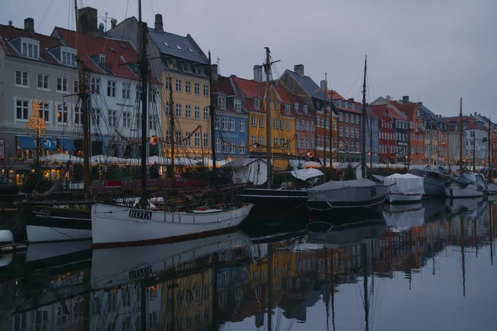 A view of a canal in a city with several colorful buildings with orange yellow and blue walls and many sailboats parked in the water under an overcast sky