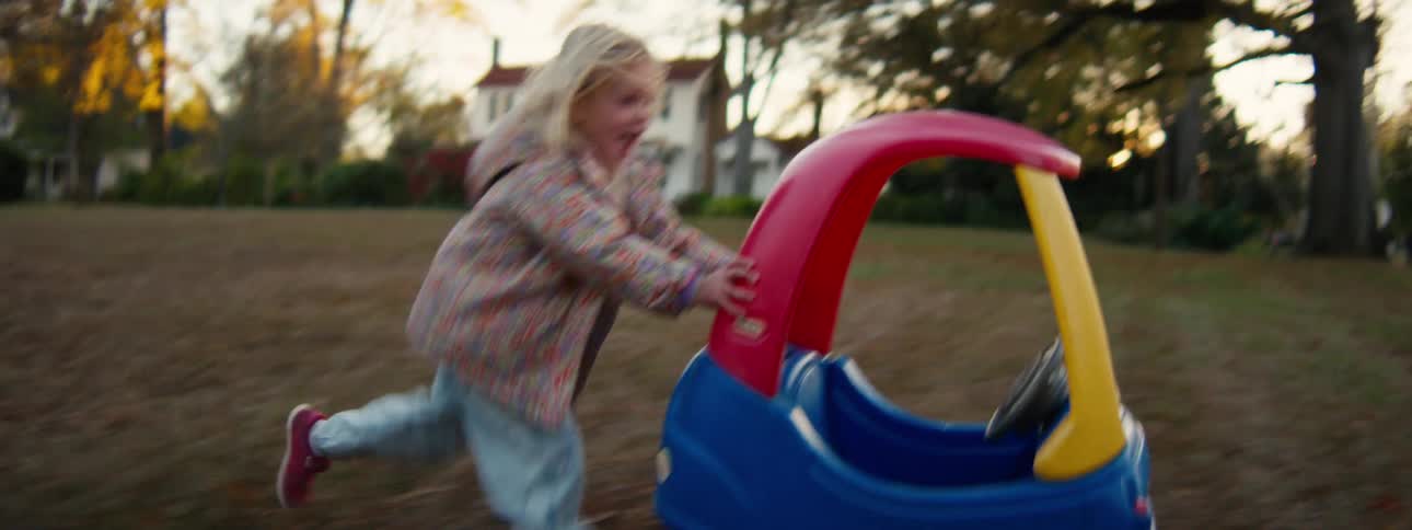 Young girl pushing a colorful toy car on a grassy area with trees and a house in the background
