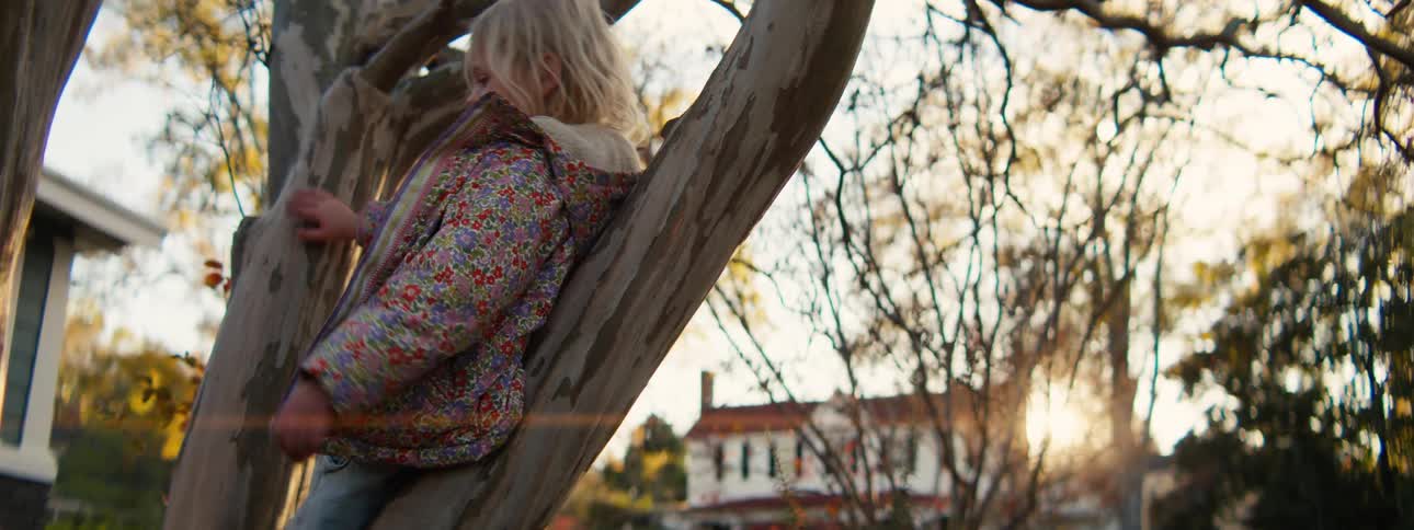 Child wearing a floral jacket climbs a tree trunk in a residential area during autumn with lowangle sunlight
