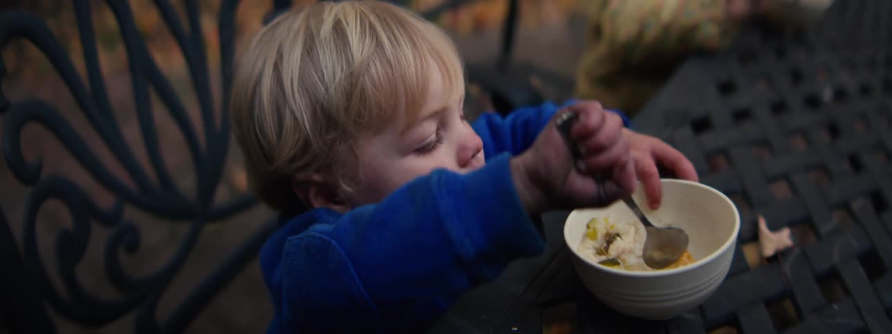 A young blond boy in a blue sweater sits at an outdoor table and attempts to eat from a white bowl he uses both a spoon and his hands to handle the food before looking toward the camera with a frustrated or upset expression