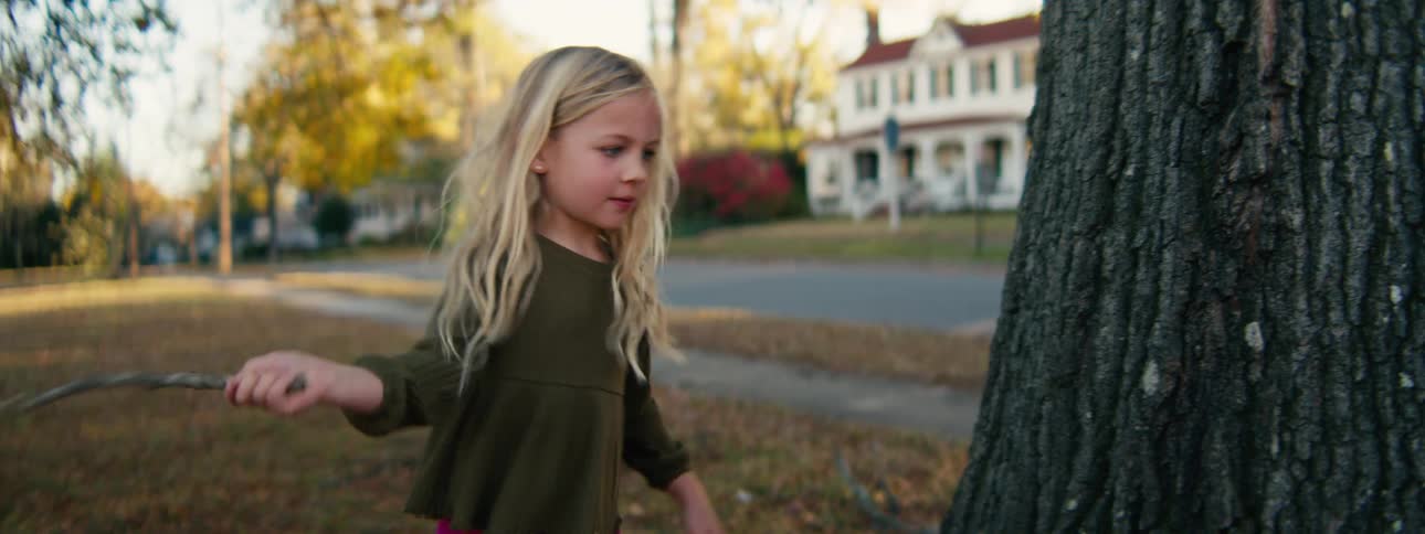 A young girl angrily hits a tree trunk with a stick in a quiet suburban yard