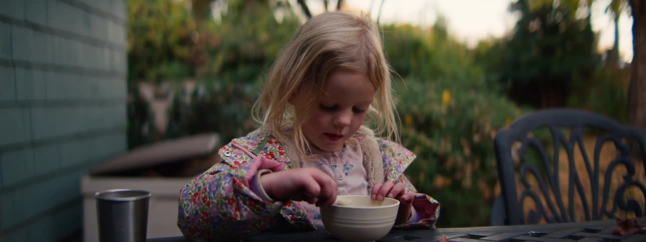 A young girl eats from a small bowl at a table outdoors in a peaceful backyard