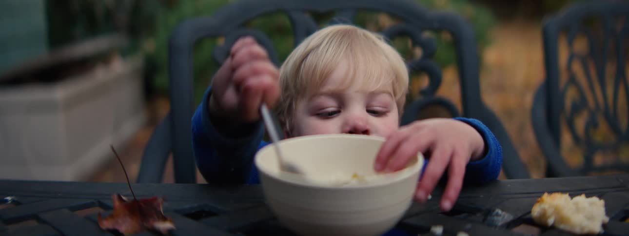 A young boy with blonde hair sits at an outdoor table and uses a spoon to eat from a large white bowl
