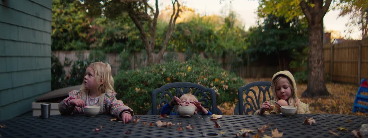 Three young children sit at a table eating from bowls amid colorful autumn leaves in a backyard