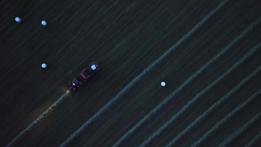 A highangle drone shot shows a tractor with headlights on pulling a baler through a dark field at night leaving behind white wrapped hay bales while following diagonal rows of cut grass across the vast agricultural landscape
