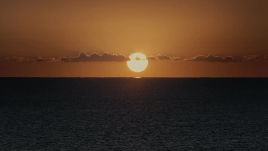 A bright sun descends toward the horizon of a calm ocean under a deep orange sky with a thin layer of clouds