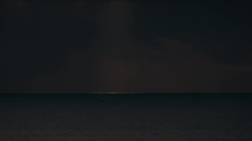 A distant wide shot shows a thin horizontal line of golden city lights shimmering on the horizon across a dark calm body of water at night