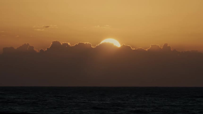 The sun begins to set behind a thick bank of dark clouds over a vast calm ocean the entire sky is filled with a deep golden and orange glow as the last light of the day disappears