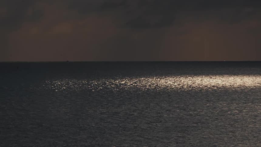 A wide shot shows a calm body of water at dusk with sunlight reflecting in a shimmering patch across the center of the surface under a dark heavy sky