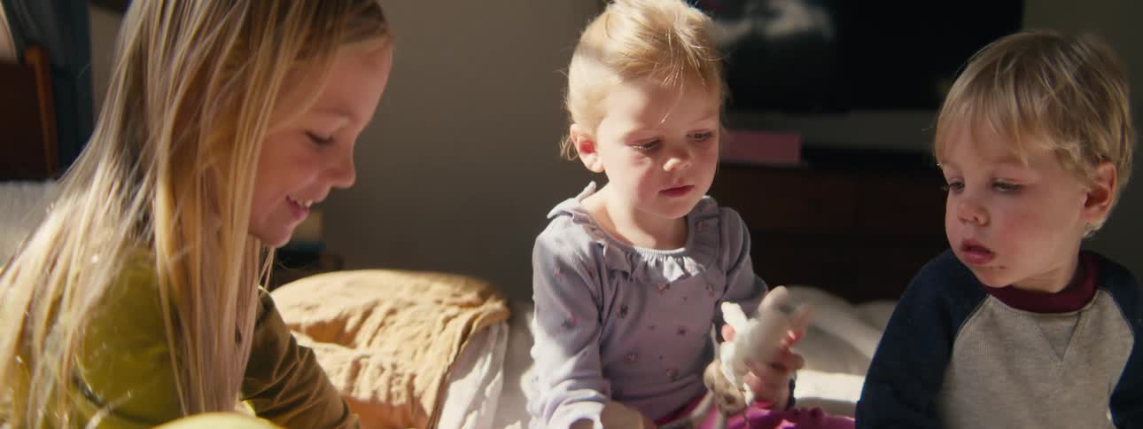 Three young children playing with toys together on a bed in a softly lit bedroom