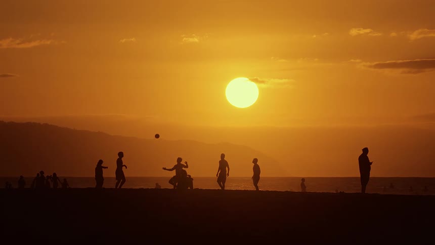 Silhouetted people gather and play along the beach as the sun sets over the ocean at dusk