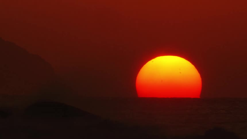 The sun glows on the horizon in a deep red sky as waves roll in to shore
