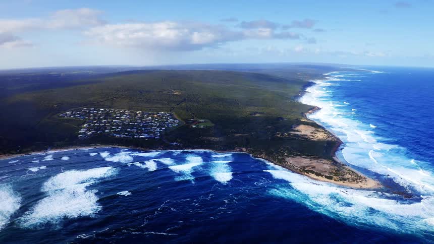 Aerial view of a rugged coastline with crashing ocean waves and green land extending into the sea