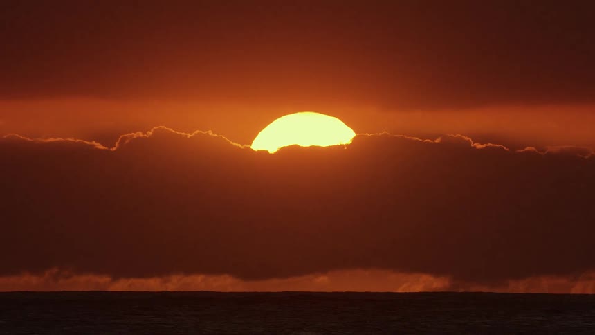 A large sun slowly descends behind a thick layer of dark clouds over the ocean casting a deep orange glow across the sky and reflecting off the waters surface until the sun disappears completely behind the lower cloud bank