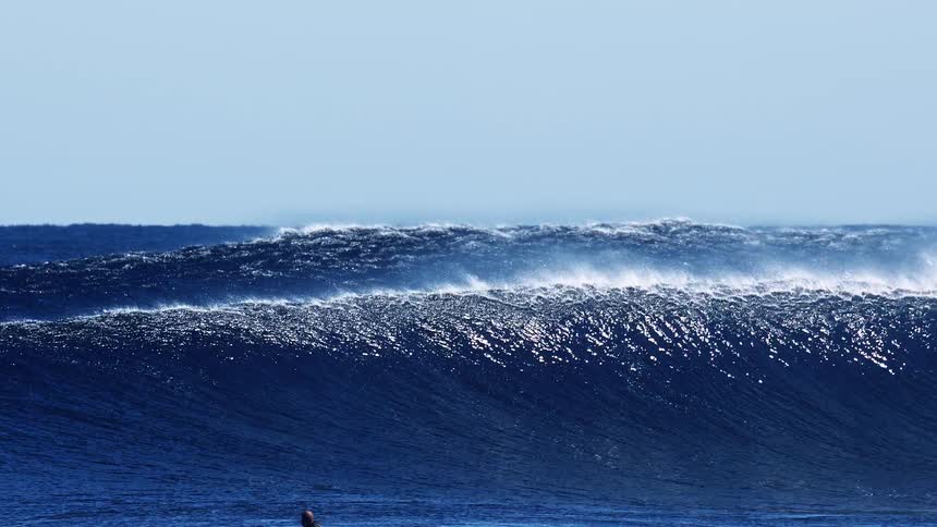 Large dark blue ocean swells roll forward and crest under a clear sky creating white sea spray and mist as the heavy waves begin to break across the horizon in open water