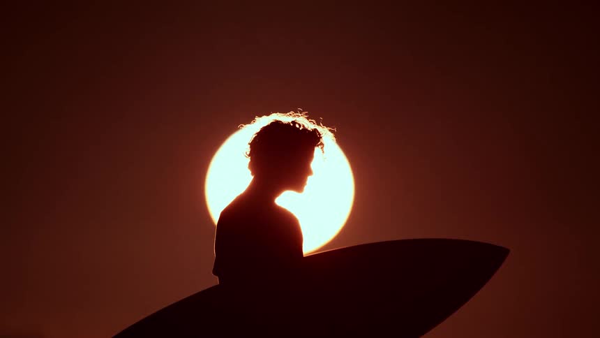 A silhouette of a male surfer holding his surfboard standing against a large bright setting sun