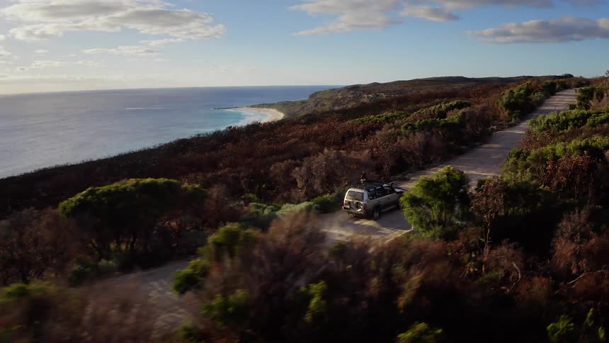 Aerial view of a vehicle driving along a winding coastal road by the ocean