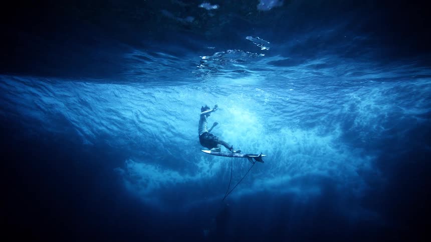 A male surfer performs a duck dive beneath a large breaking wave pushing his board under the turbulent white water while sunlight filters through the deep blue ocean surface from an underwater perspective