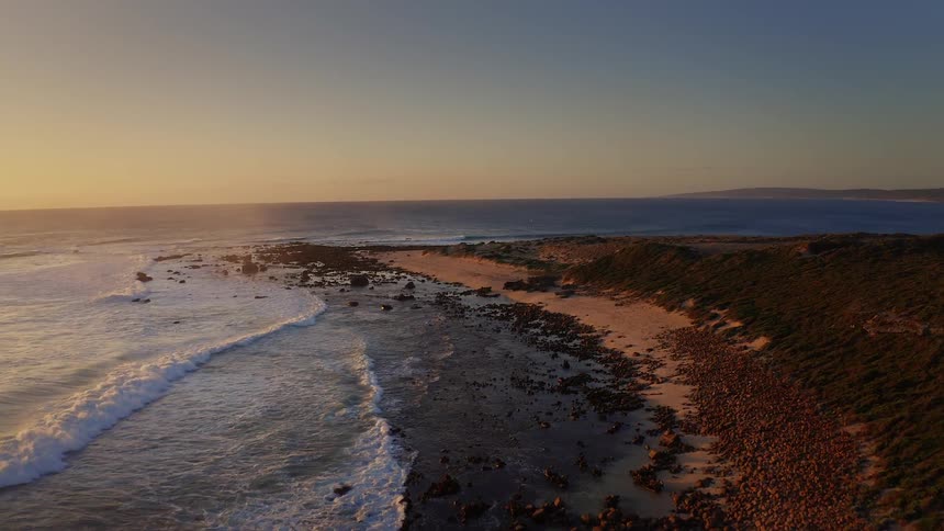 This drone footage tracks along a rugged shoreline at sunset moving over a dense field of brown rocks toward a sandy beach where waves crash against the coast and sunlight reflects off the ocean surface beneath a clear orange sky