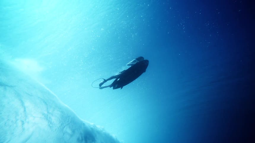 A surfer is seen from an underwater perspective performing a duck dive to push their surfboard beneath a large breaking wave the shot captures the bright sunlight filtering through the clear blue water as bubbles swirl around the athlete and their board