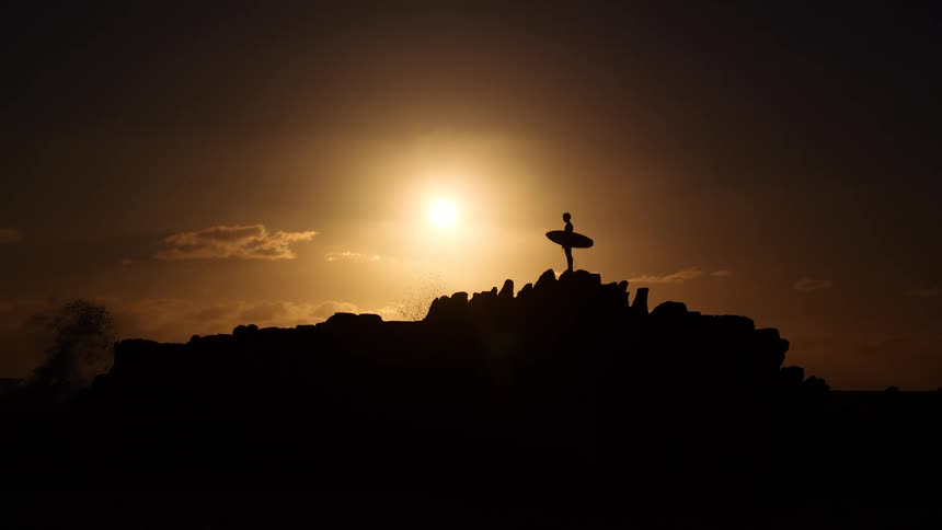A surfer stands atop a rocky cliff silhouetted against a bright golden sun low in the sky the dark outline of their surfboard is clearly visible as they look out over the horizon during the sunset