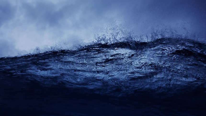 A dark powerful ocean wave crests and breaks against a moody overcast sky the camera then plunges beneath the surface capturing the turbulent blue water and rising air bubbles from an underwater perspective