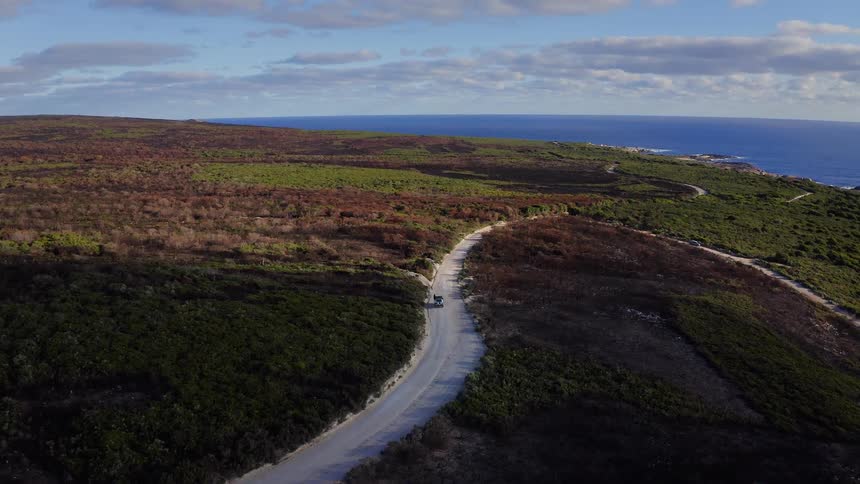 Aerial view of a vehicle traveling along a winding coastal dirt road leading to the ocean