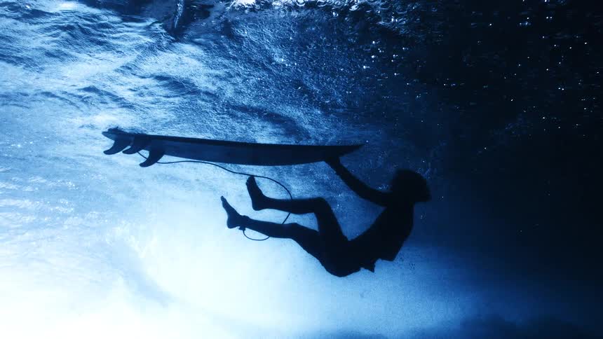 A silhouette of a surfer pulls their board downward to duck dive under a large breaking wave viewed from an underwater perspective as white foam and turbulent water churn above them in the deep blue ocean