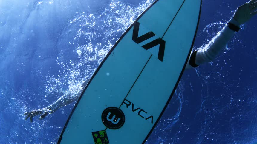 A male surfer paddles through clear blue water as seen from directly underneath the surfboard showing his arms pulling through the water and a trail of white bubbles against the bright sunlight filtering down from the surface