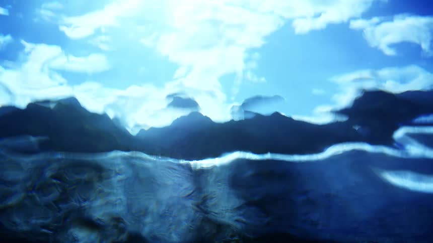Wave breaking overhead obscures the cloudy sky and coastal mountains visible through the water on a sunny day