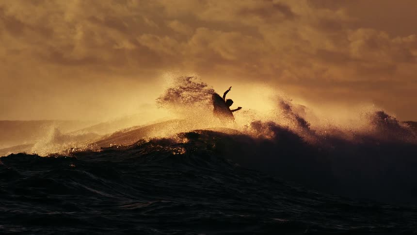 Elevated ocean wave with spray illuminated by golden light under a cloudy sky